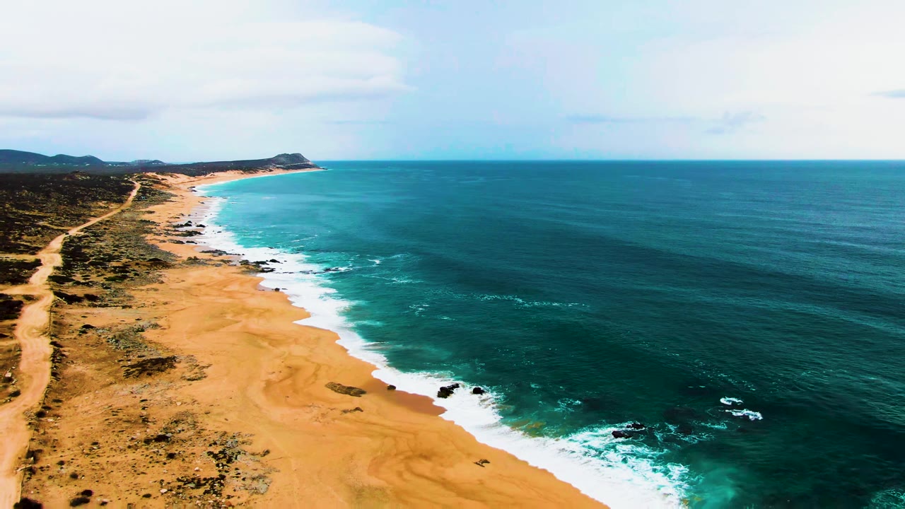 Opening aerial frame over Buzzards Beach in Baja California Sur, with sandy shore at left, white waves along the edge, deep blue ocean at right, and no visible people.