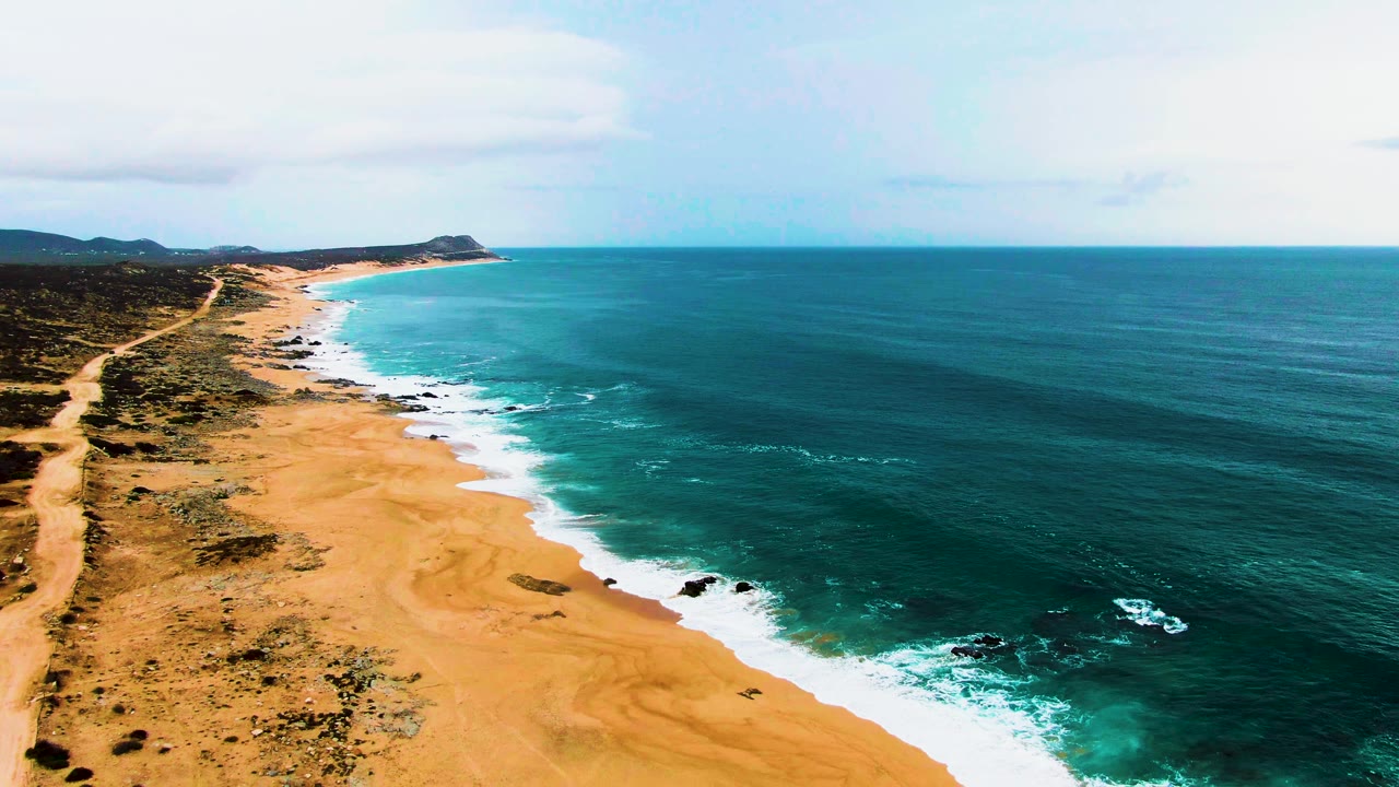 Aerial view along Buzzards Beach with the shoreline curving into the distance, turquoise water near shore, darker ocean offshore, scrubland inland, and no visible people.