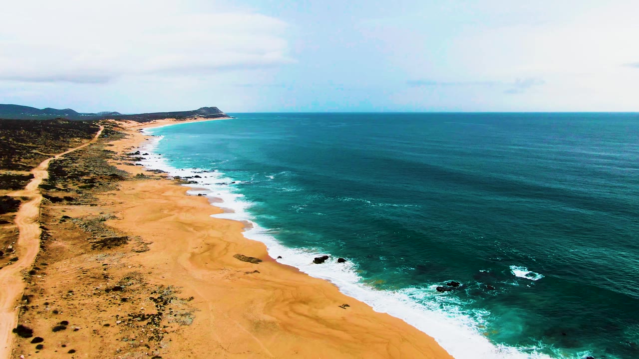 Wide aerial frame of Buzzards Beach showing a broad band of sand, white surf advancing along the shore, dark rocks in the water, inland scrub, and no visible people.