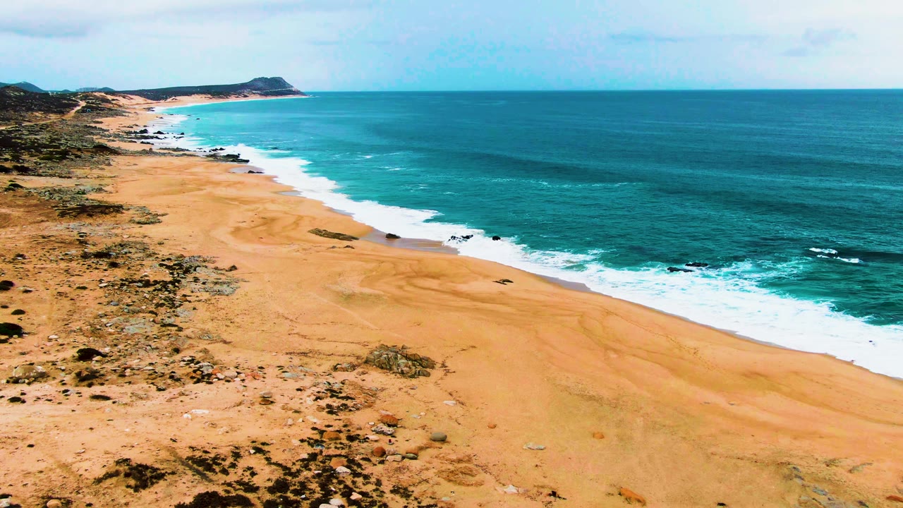 Aerial view over Punta Buzzards beach in Baja California Sur, with tan sand, sparse scrub, turquoise surf, dark shoreline rocks, a distant headland, and no visible people.