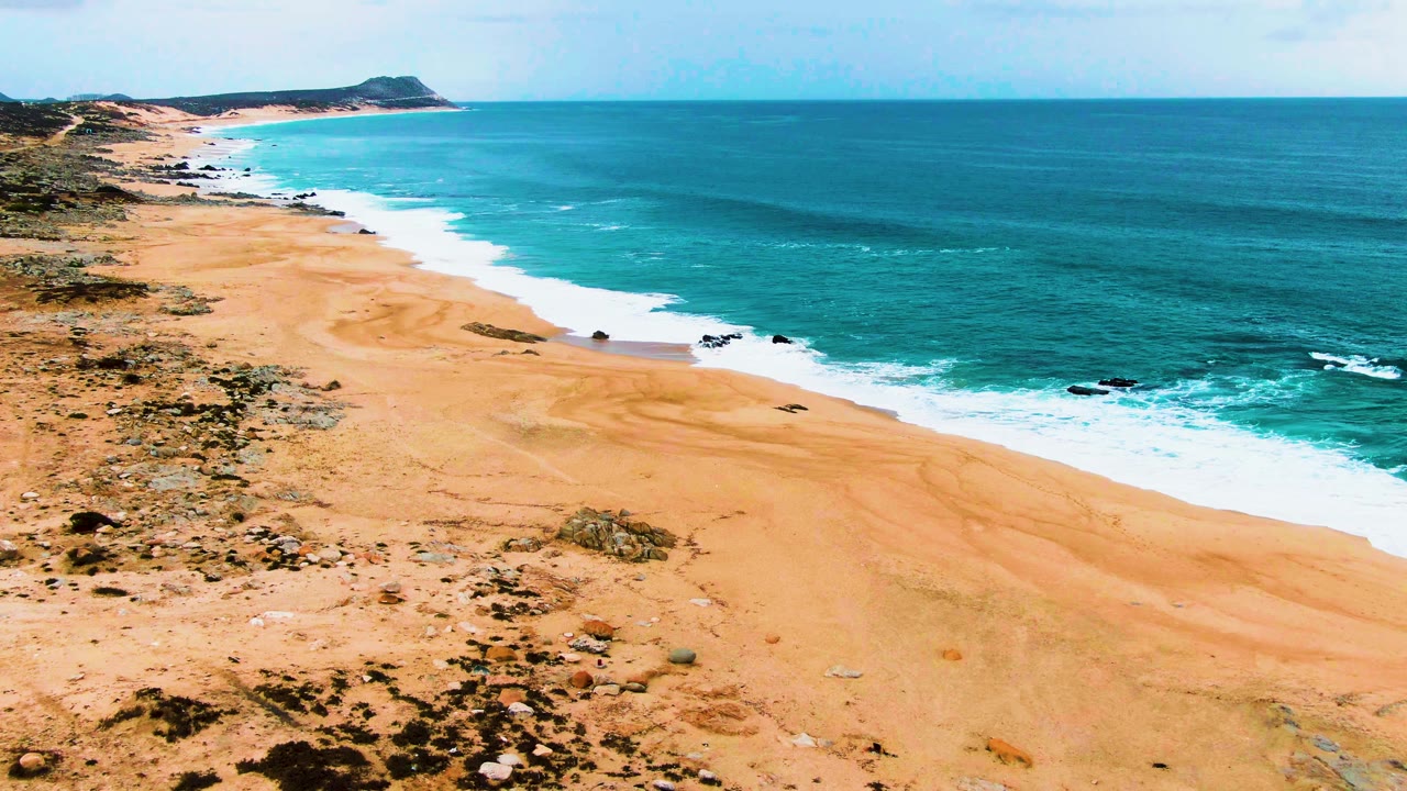 Wide coast view with tan sand, scrub, and white waves breaking near rocks.