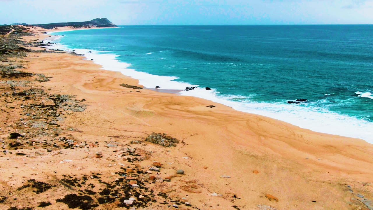 Aerial frame of Buzzards Beach on the East Cape, with orange sand, scattered scrub, white surf bands, turquoise water, a low headland, and no visible people.