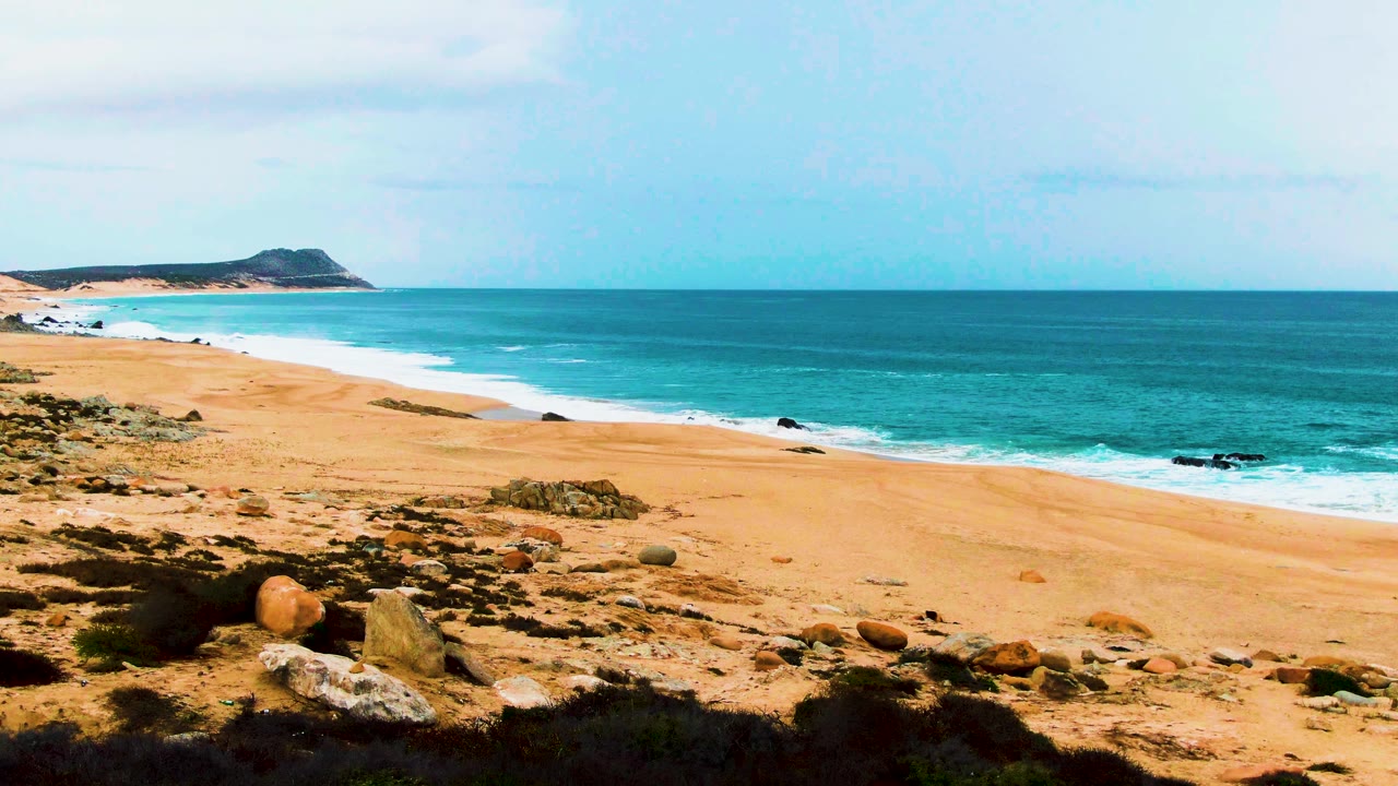 Buzzards Beach coastline in Baja California Sur with white surf tracing the sloped sand, a distant headland at left, foreground rocks, and no visible people.