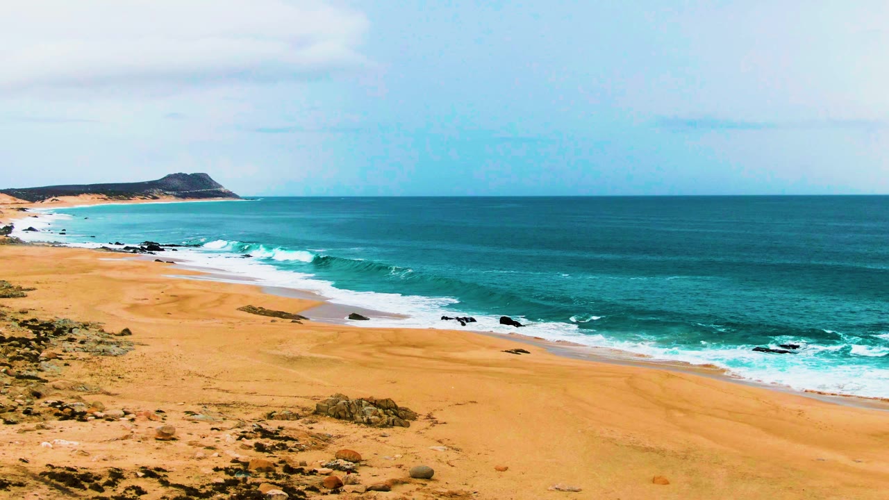 Elevated view of the Punta Buzzards beach area, with white surf bands moving toward empty golden sand, scattered dark rocks near the water, and no visible people.