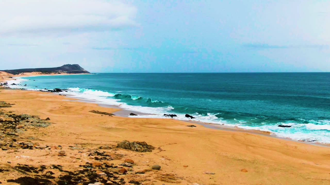 Buzzards Beach East Cape coast viewed from above, showing rocky golden sand in the foreground, a diagonal line of white foam, blue-green ocean, and no visible people.