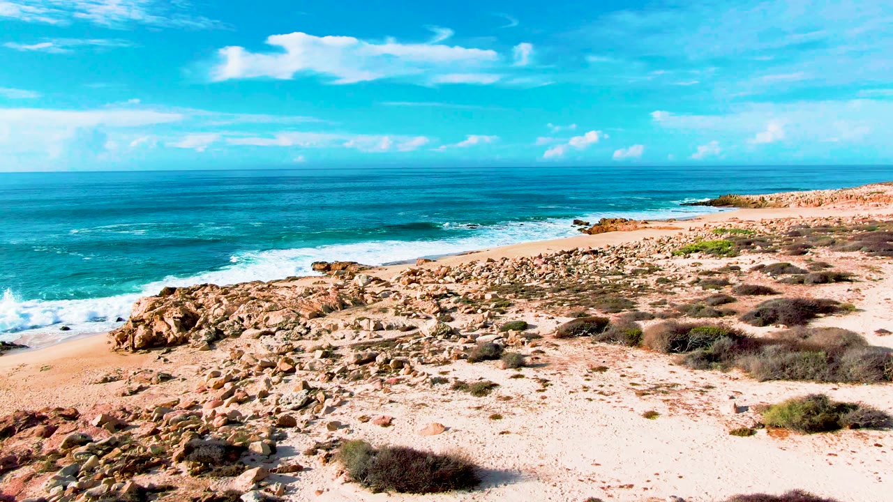 Elevated view of Punta Buzzards shoreline with rust-colored rocks, pale sand, turquoise water, white surf, blue sky, and no visible people.