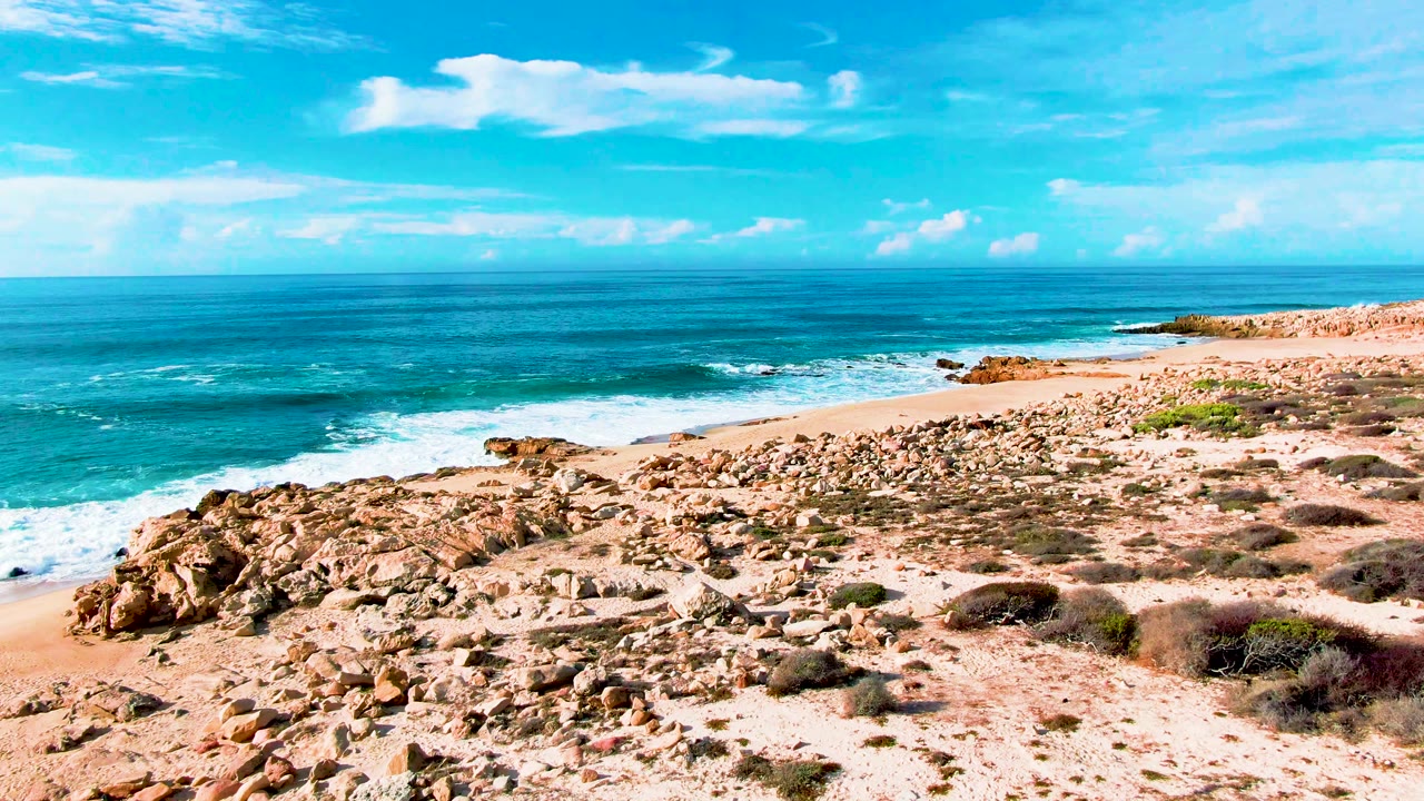 Punta Buzzards beach view with a broad rocky foreground, foaming waves along the shoreline, open blue water, scattered clouds, and no visible people.