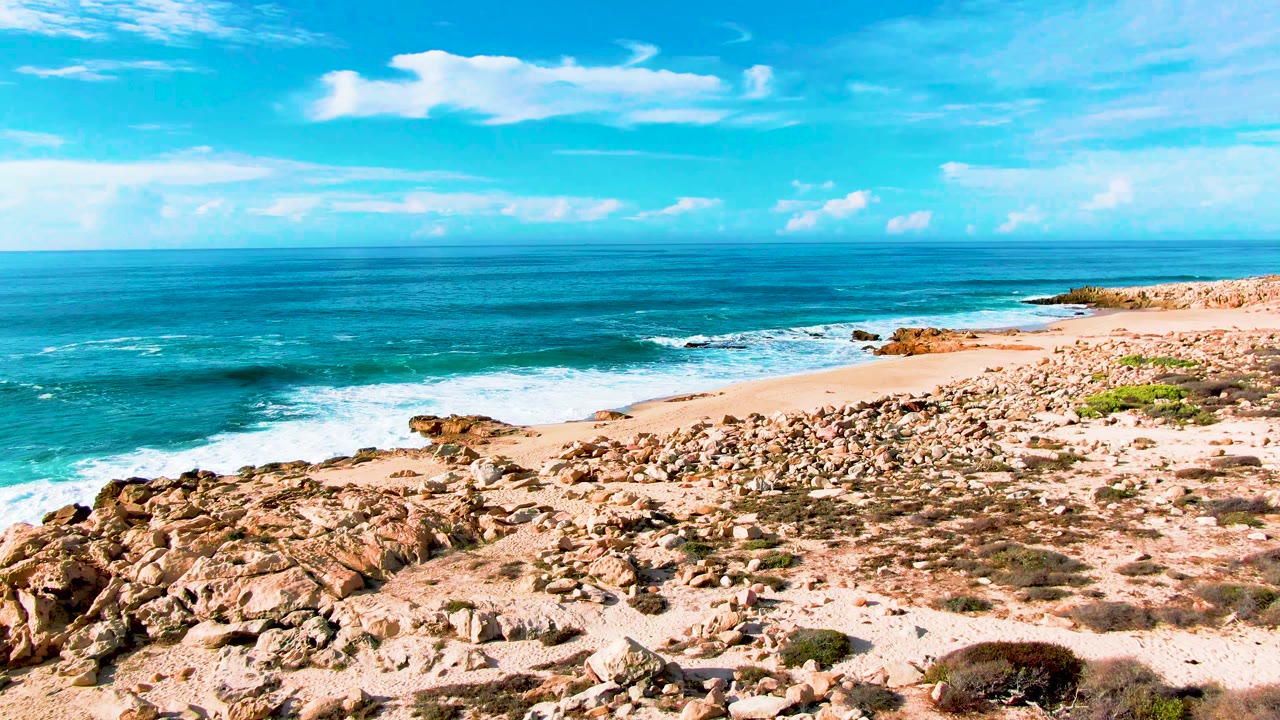 Elevated Punta Buzzards shoreline frame with a sandy cove at right, rock clusters across the beach, white wave foam at left, clear horizon, and no visible people.