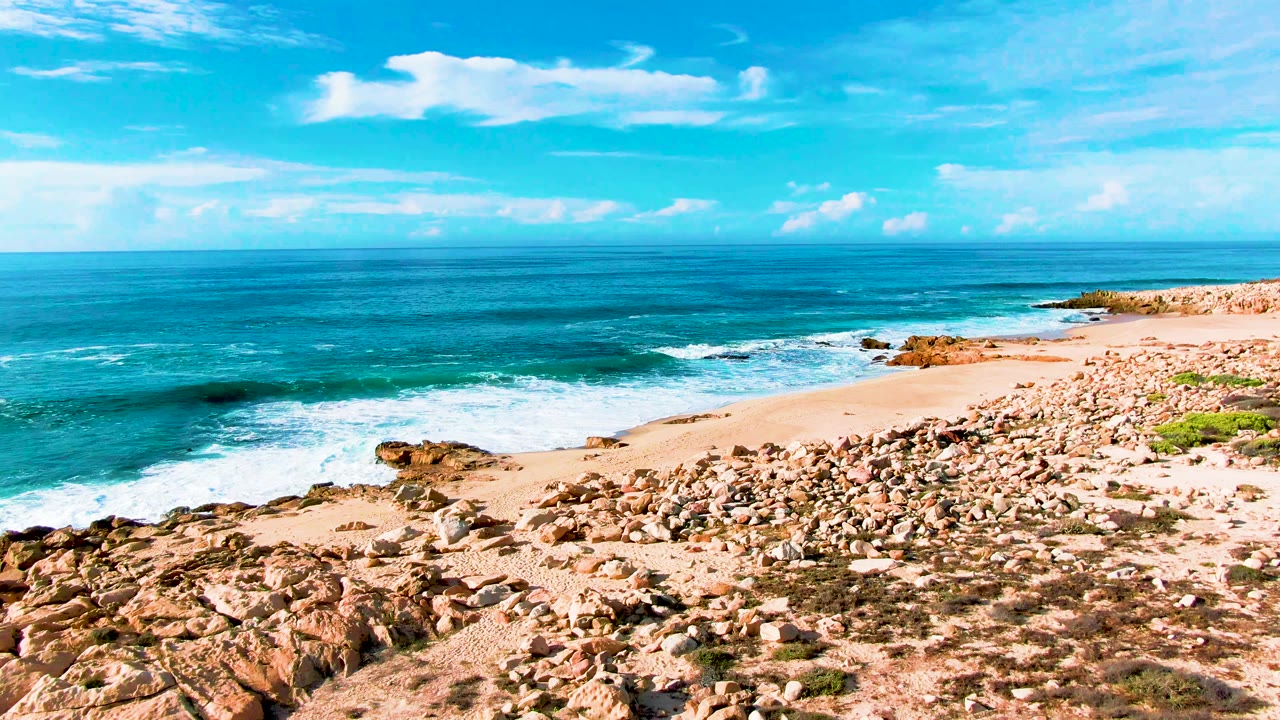 Wide coastal view with ocean, surf, rocks, sand, and blue sky