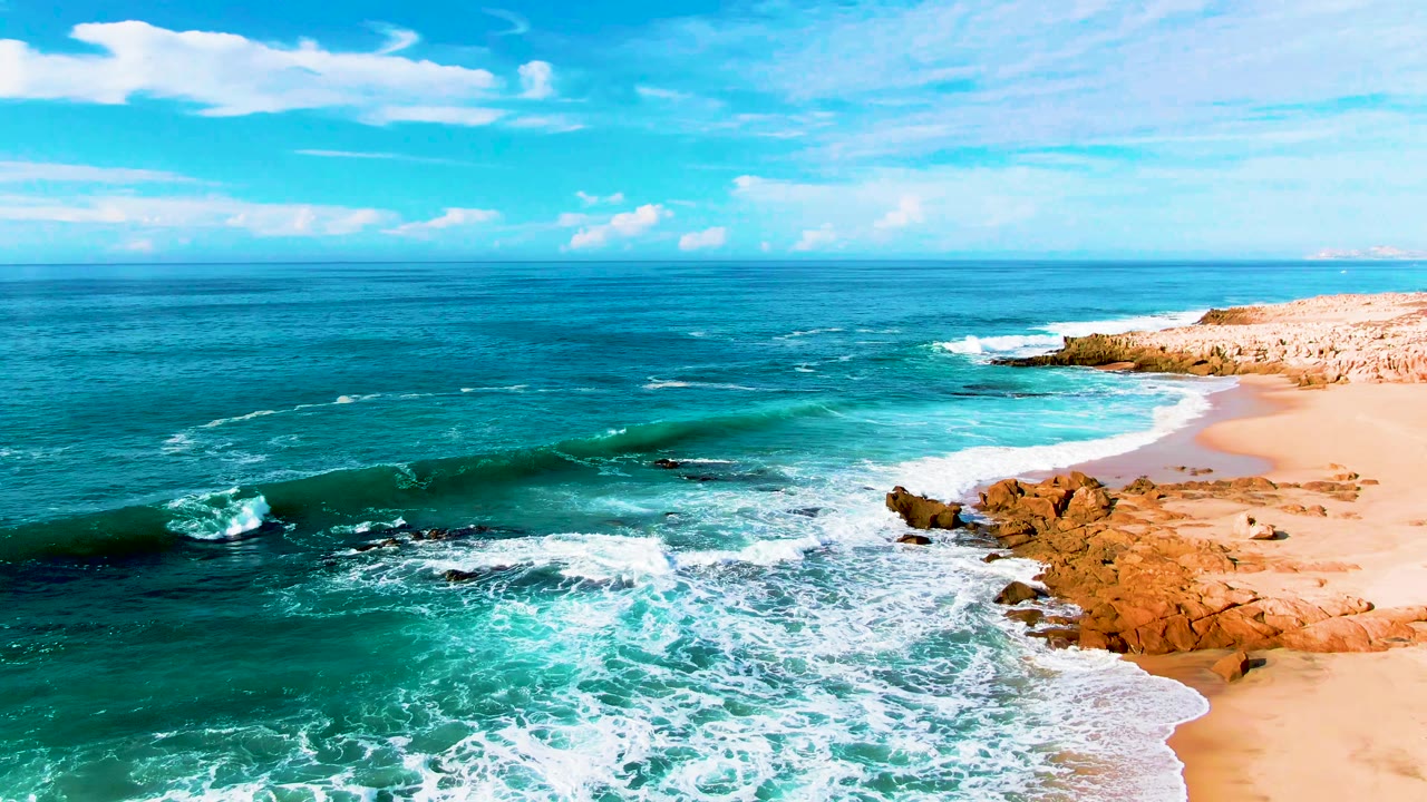 Aerial view of Punta Buzzards surf breaking over dark rocks beside a sandy beach, with turquoise water, blue sky, light clouds, and no visible people.