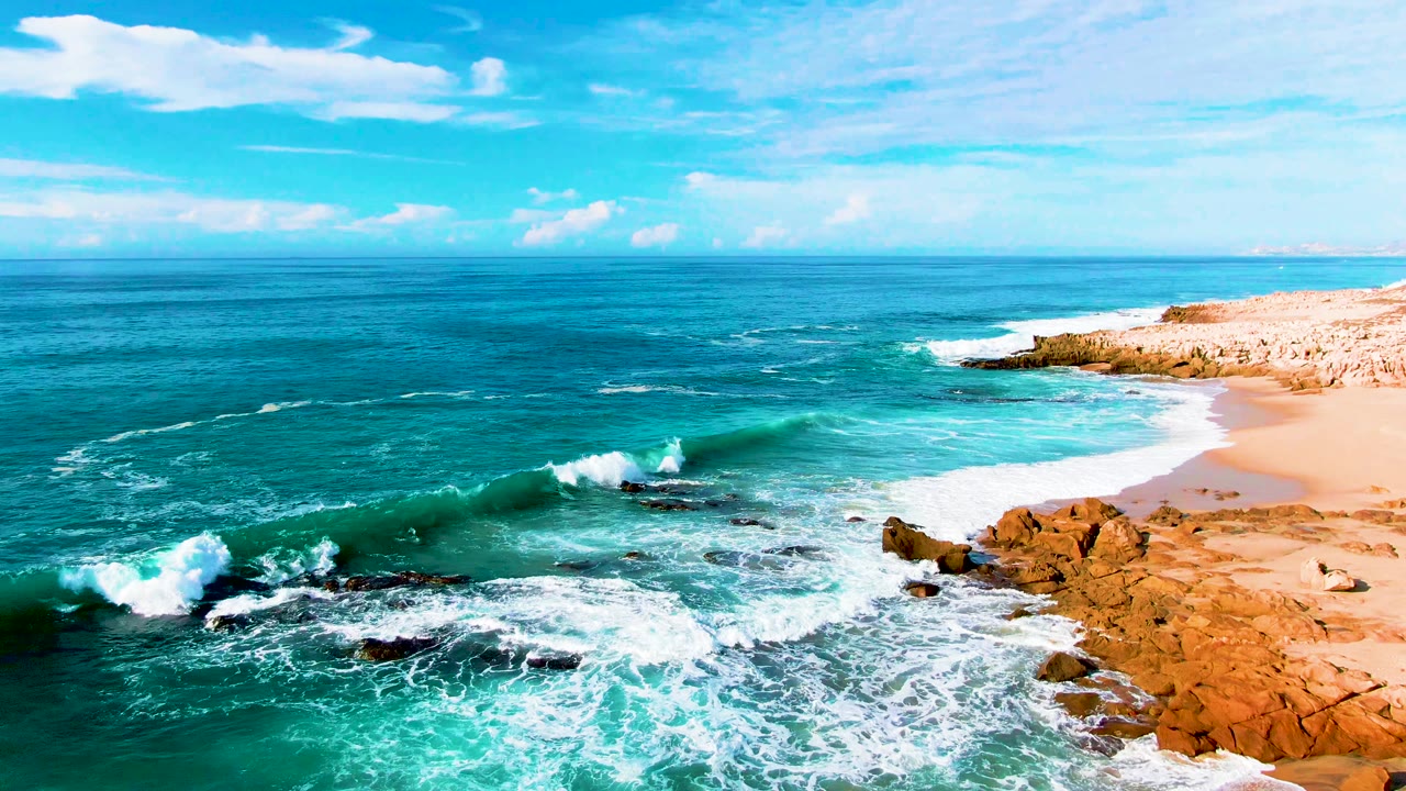 A curling turquoise wave approaches the rocky shallows at Punta Buzzards, with white foam already spread across the beach and no visible people.