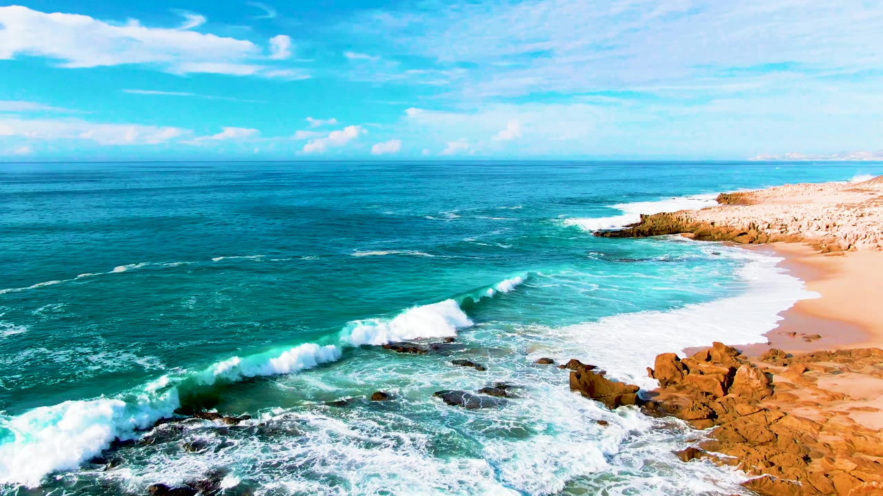 White surf breaks across the rocks at Punta Buzzards while the sandy beach curves along the right side under a clear blue sky, with no visible people.