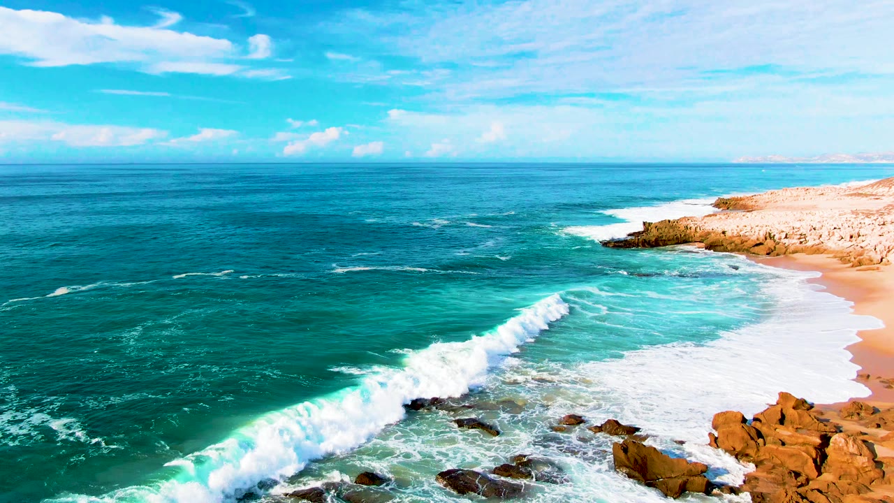 Aerial frame of Punta Buzzards with a long foam line sweeping along the sandy beach, dark rocks in the lower frame, and no visible people.
