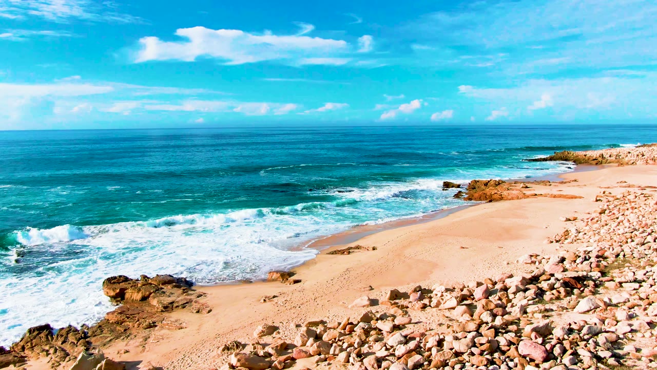 At Punta Buzzards in Baja California Sur, turquoise surf approaches a tan sandy beach with dark rocks in the foreground and no visible people.