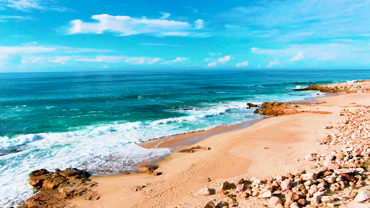 At Punta Buzzards in Baja California Sur, white foam spreads farther across the sand beside a pebble-covered beach edge, with no visible people.