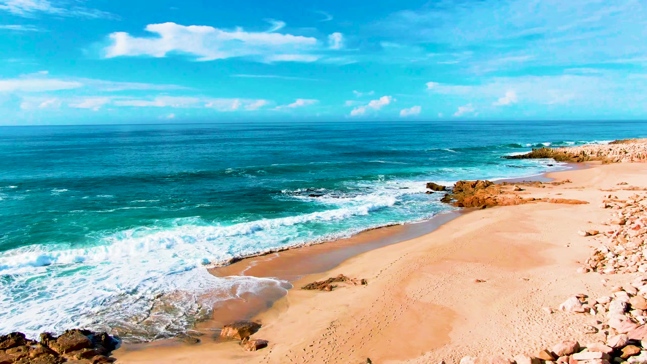 At Punta Buzzards in Baja California Sur, a breaking wave curls into a sandy cove bordered by rocks, with an open blue horizon and no visible people.