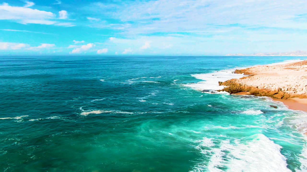 At Punta Buzzards in Baja California Sur, turquoise surf rolls toward a pale beach and rocky shoreline under a partly cloudy sky, with no visible people.