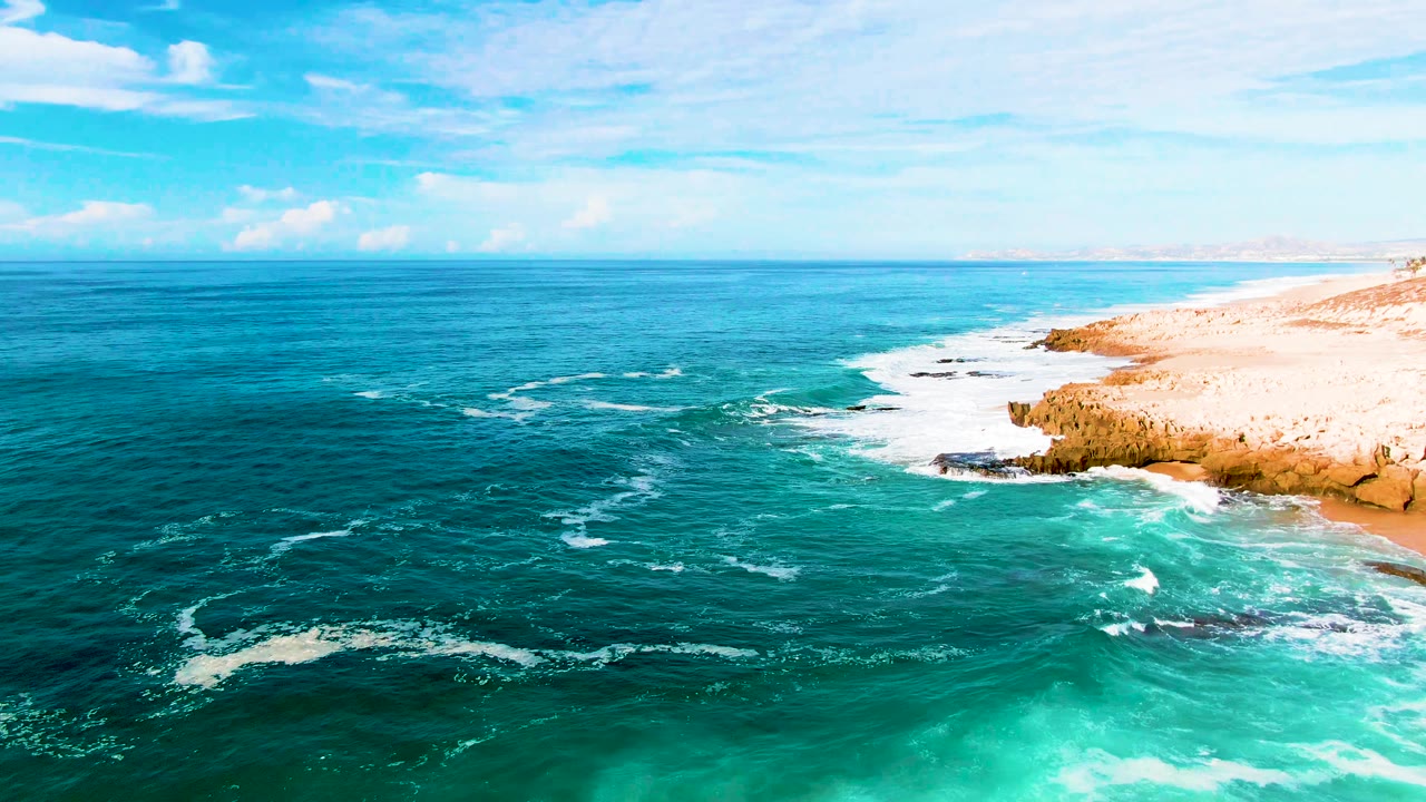 Deep blue water dominates the frame while scattered foam lines approach the rocky East Cape shore and pale beach at Punta Buzzards, with no visible people.