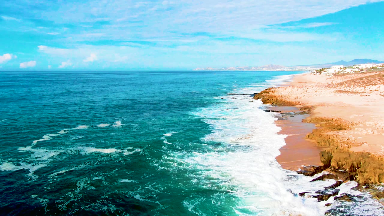 Aerial frame from Buzzards Beach, East Cape, Baja California Sur, showing turquoise water, white surf, sandy shoreline, rocky edges, distant coast, and no visible people.