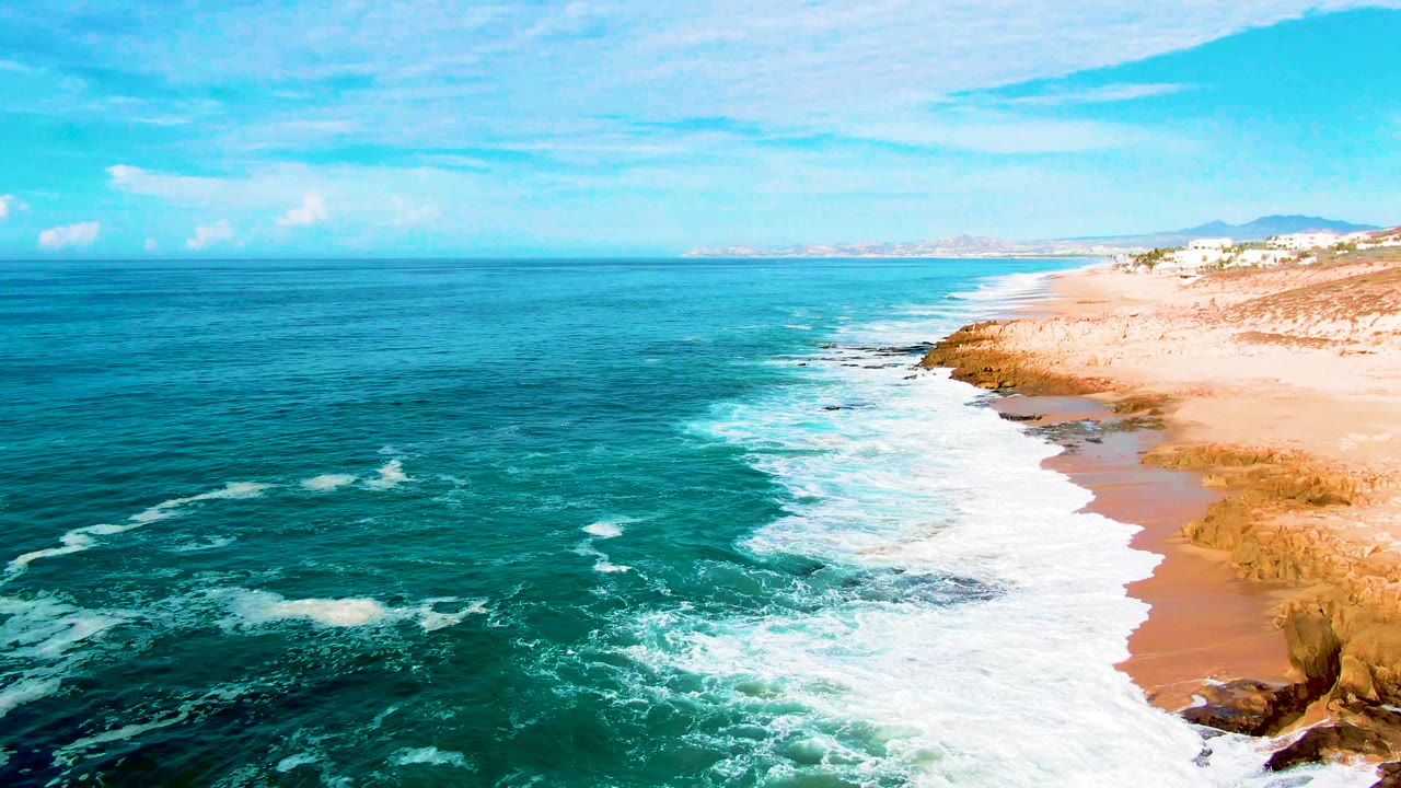 White ocean foam spreads across a sandy beach beside rocky shoreline.