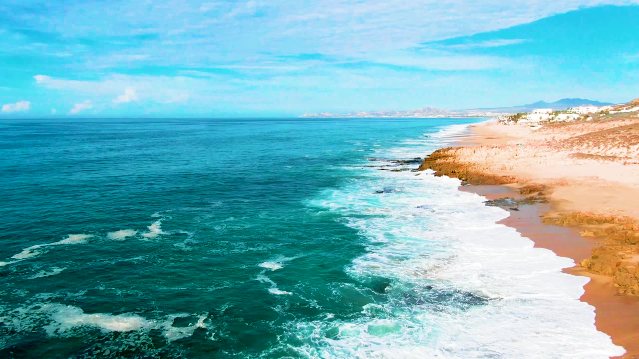 Aerial view at Buzzards Beach, East Cape, Baja California Sur, where broad whitewater covers the near shore, turquoise water remains offshore, rocks break the beach edge, and no visible people appear.