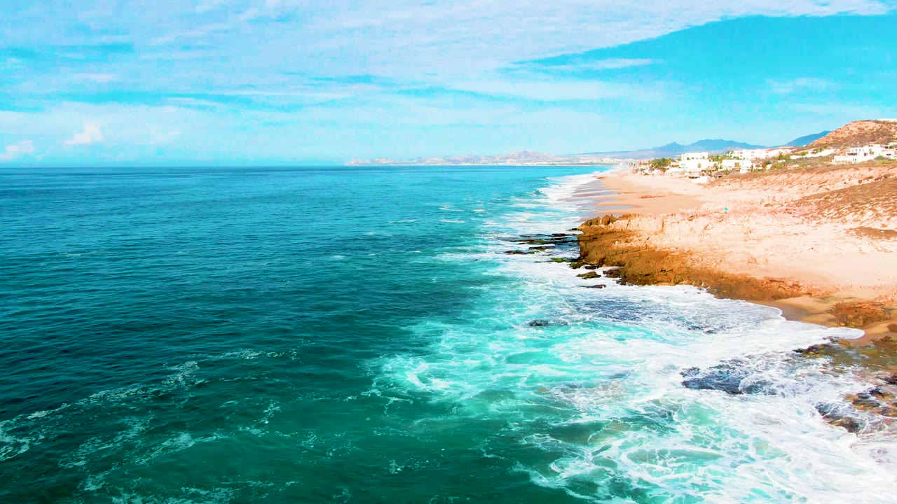 Aerial view at Punta Buzzards, Baja California Sur, with turquoise surf curling over dark rocks beside a sandy beach and low hills in the distance.