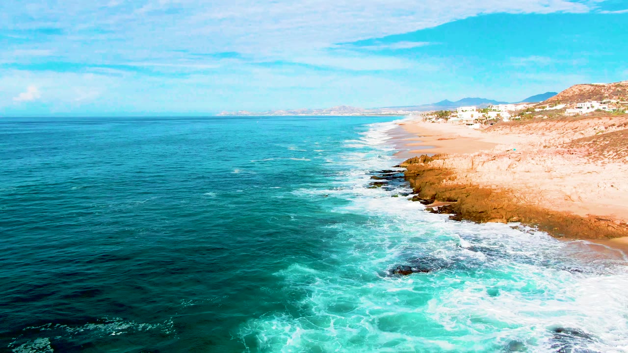 Punta Buzzards shoreline frame with white foam sweeping around a rocky point, open ocean on the left, and beach and hillside structures on the right.