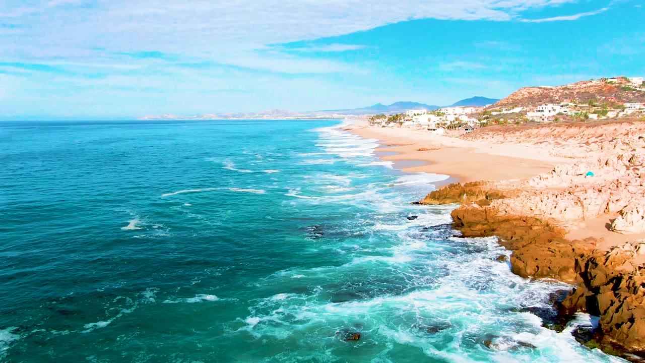 Aerial view at Punta Buzzards, Baja California Sur, with turquoise ocean on the left, white surf around dark rocks, sandy beach to the right, hillside structures in the distance, and no visible people.