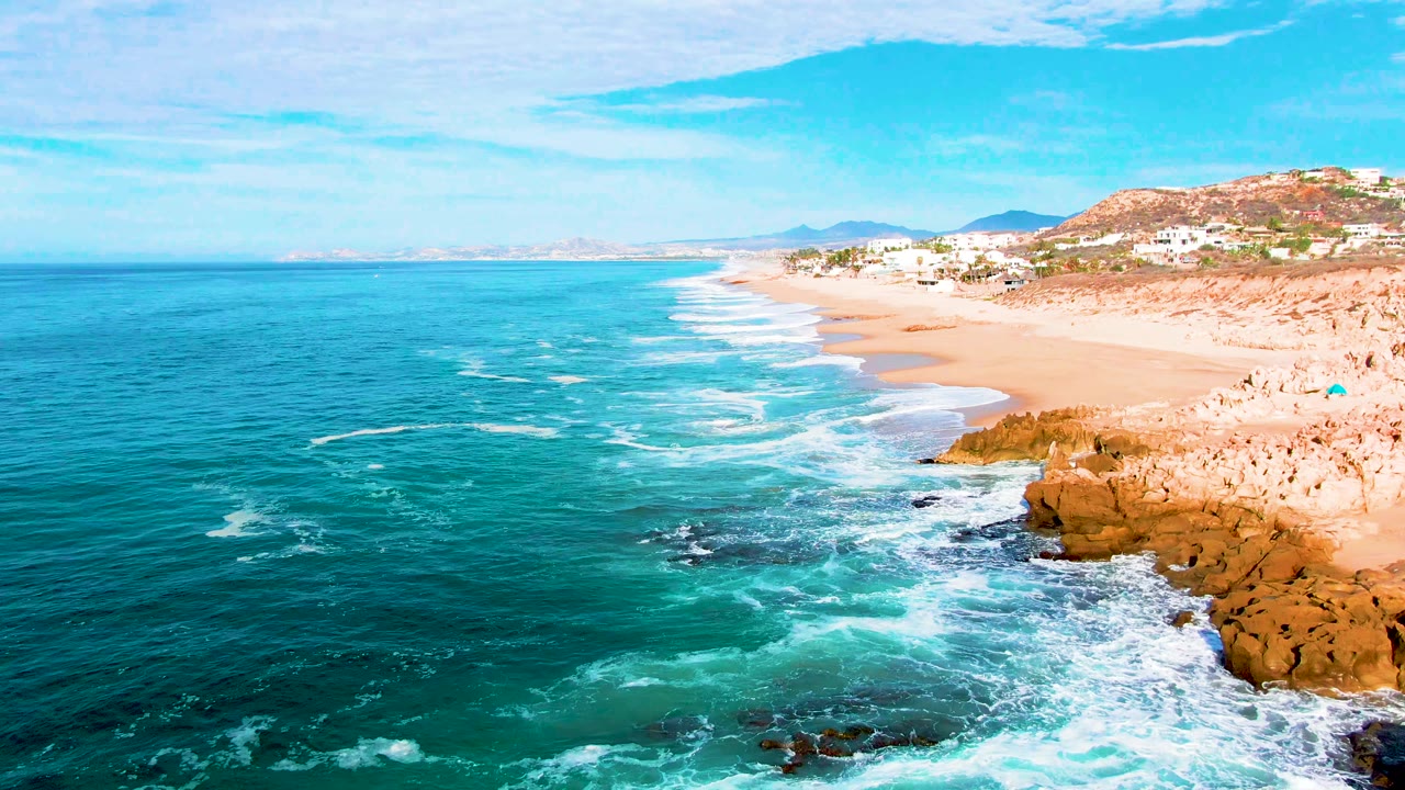Wide aerial coastline with white waves curling toward a sandy beach and rocks.
