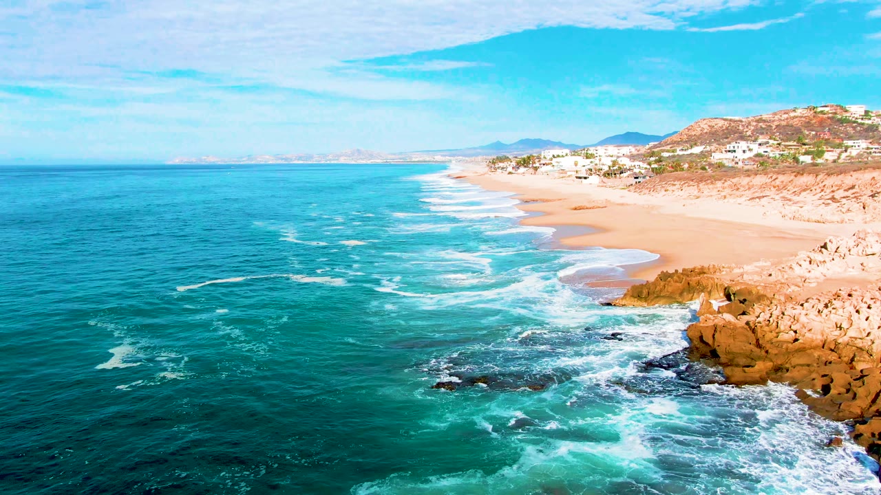 Aerial frame from Punta Buzzards, Baja California Sur, with foamy surf spreading across rocks in the lower right, open blue water to the left, a long sandy beach ahead, and no visible people.