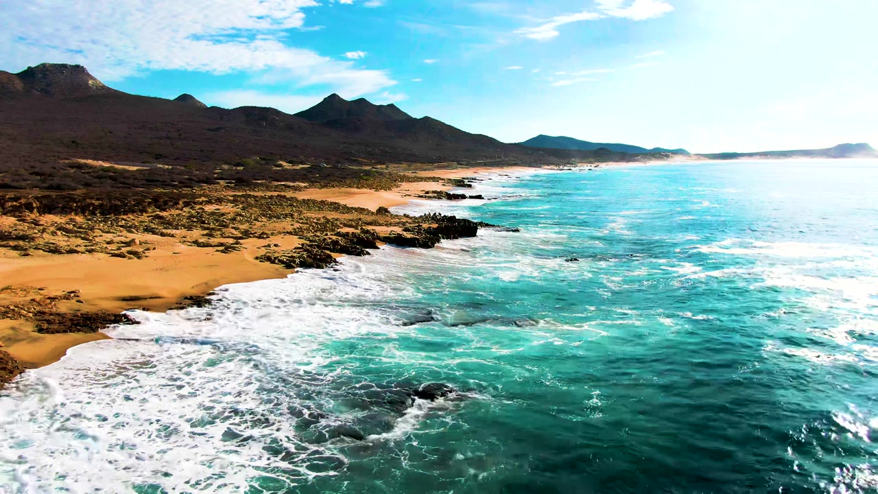 Aerial view at Punta Buzzards showing turquoise ocean on the right, white surf reaching dark rocks and orange sand on the left, mountains in the distance, and no visible people.