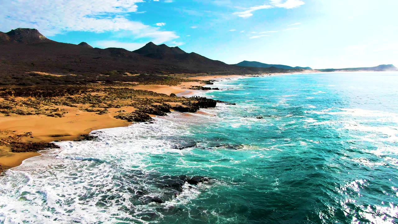 At Punta Buzzards, white foam spreads farther across the near shoreline while the turquoise water runs along the beach, with dark rocks, distant mountains, blue sky, and no visible people.