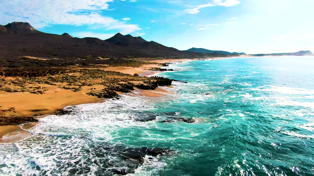 Aerial frame from Punta Buzzards with a bright band of surf curling into the sandy cove, black rocks breaking the shoreline, open ocean to the right, mountains beyond, and no visible people.