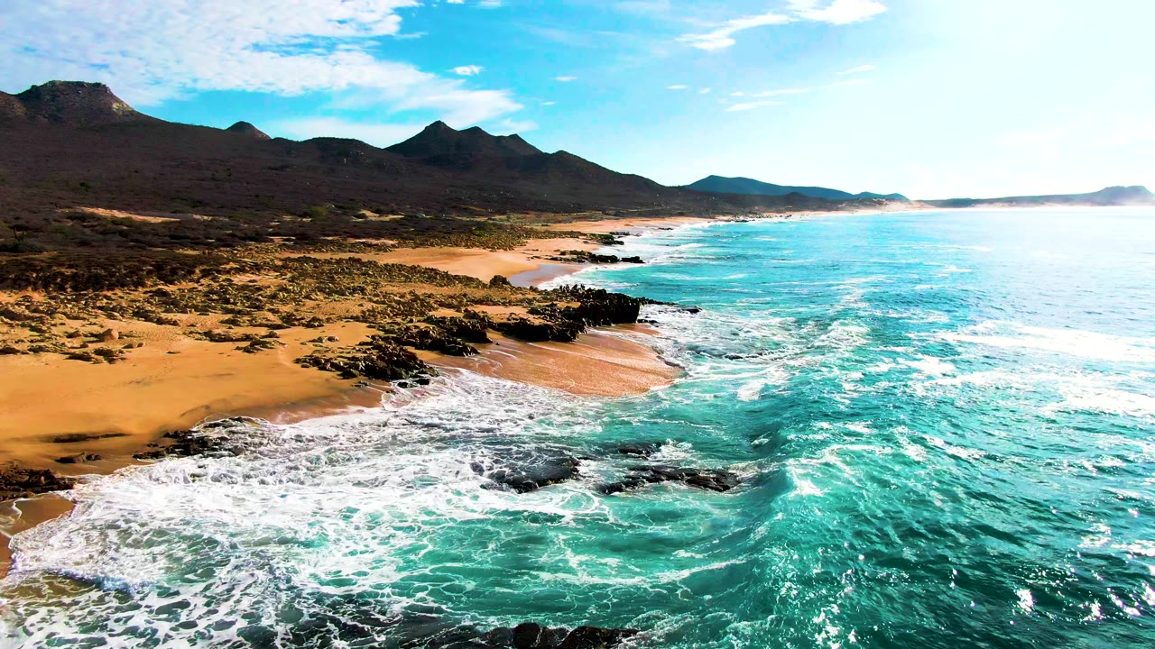 Coastal drone view of waves washing around rocks on a sandy beach.