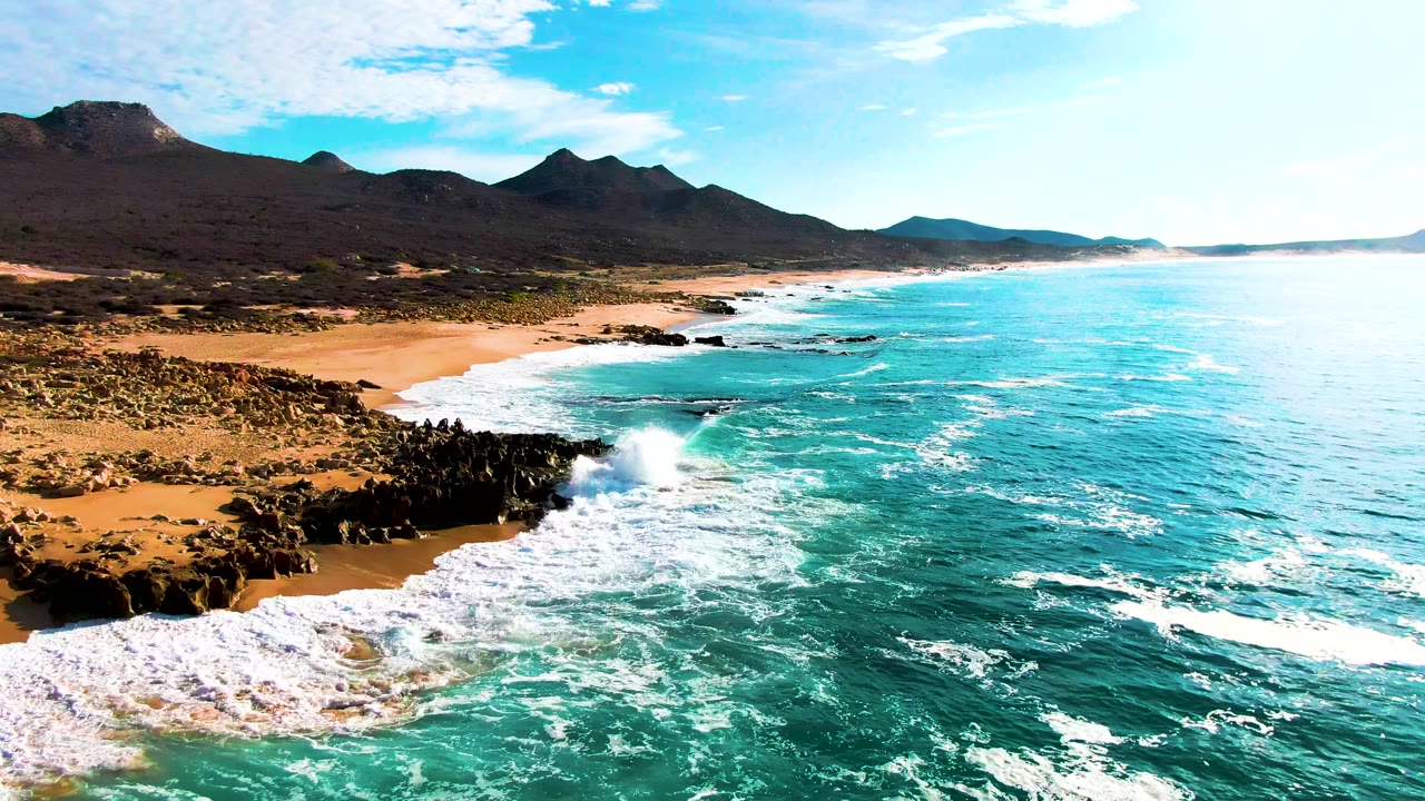 Aerial frame over Buzzards Beach in Baja California Sur, Mexico, with white surf hitting dark shoreline rocks, turquoise water to the right, sandy beach to the left, mountains in back, and no visible people.