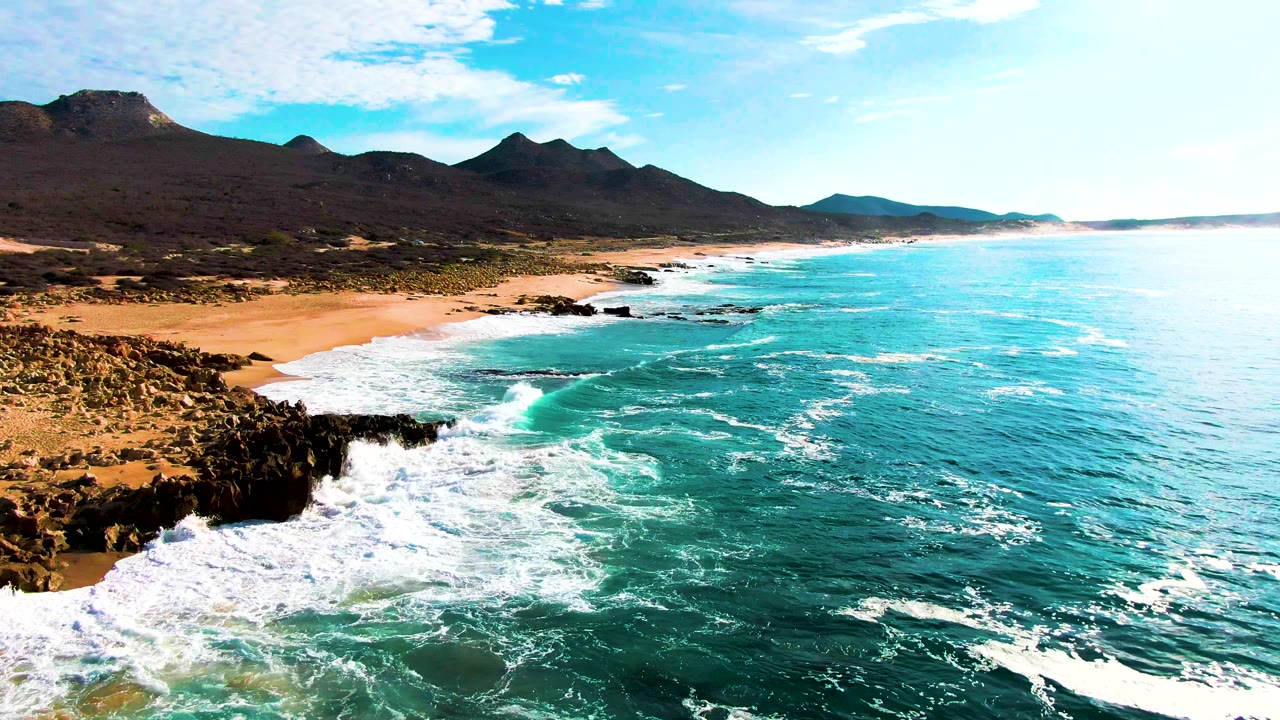 Aerial view at Buzzards Beach with surf foam widening around the foreground rocks, a sandy shoreline running into the distance, turquoise water offshore, mountain ridges inland, and no visible people.