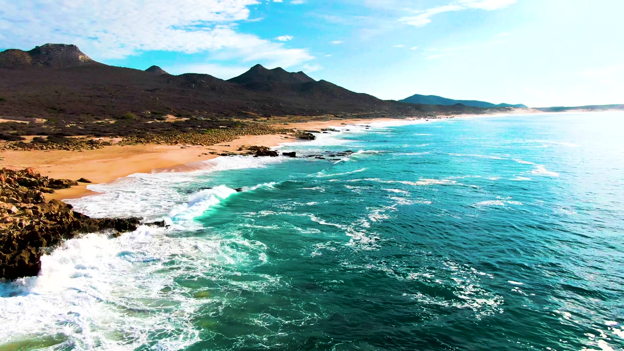 Aerial coastal frame from Buzzards Beach showing a larger expanse of turquoise ocean, foamy waves breaking near dark rocks, tan sand along the shore, distant mountains, and no visible people.