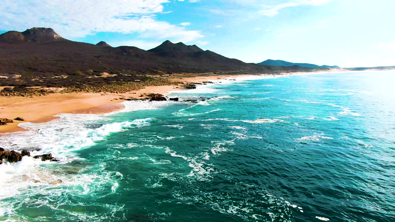 Aerial frame over Buzzards Beach with the shoreline stretching diagonally across the image, white waves washing over scattered rocks, open sand inland, mountains on the horizon, and no visible people.