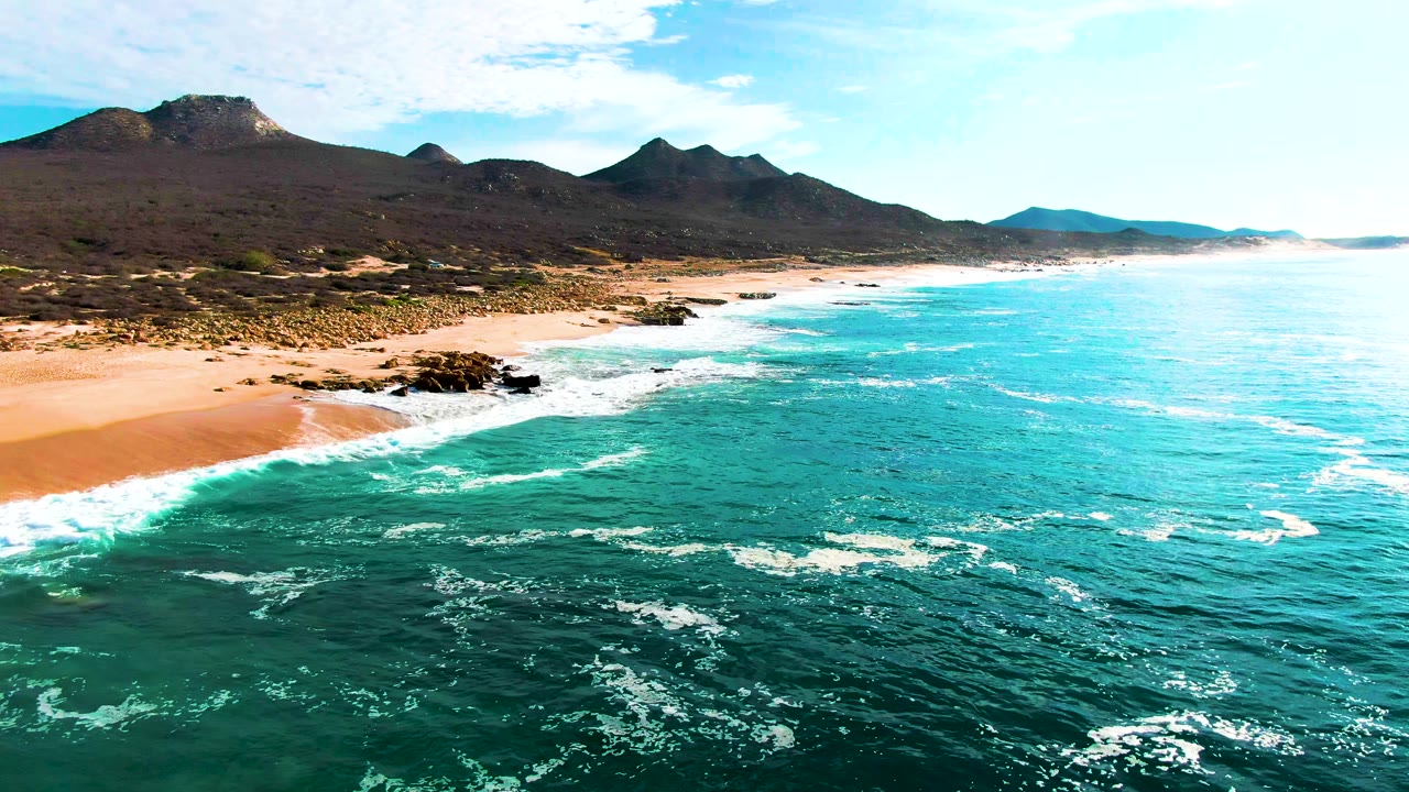 Aerial view at Punta Buzzards, Baja California Sur, with turquoise ocean in the foreground, sandy beach and dark rocks along the shore, distant mountains under a partly cloudy sky, and no visible people.