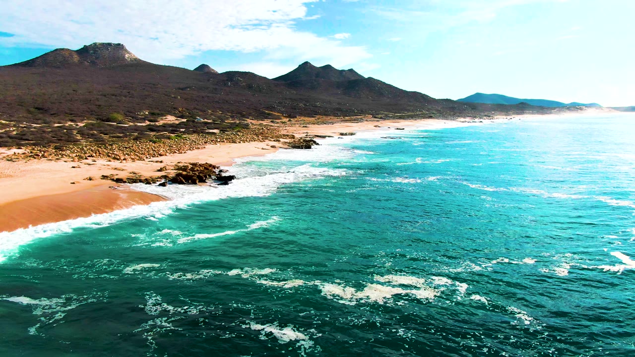 Wide aerial frame at Punta Buzzards showing white waves moving toward rocky outcrops, a pale sandy beach, dark coastal terrain, mountain ridges, and no visible people.