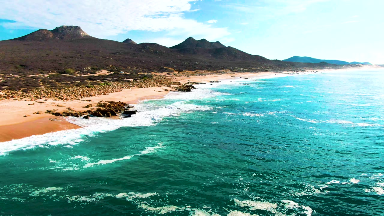 Aerial view of Punta Buzzards coastline with a broader spread of turquoise water, white foam bands near the rocks, sand along the left shore, distant mountains, and no visible people.