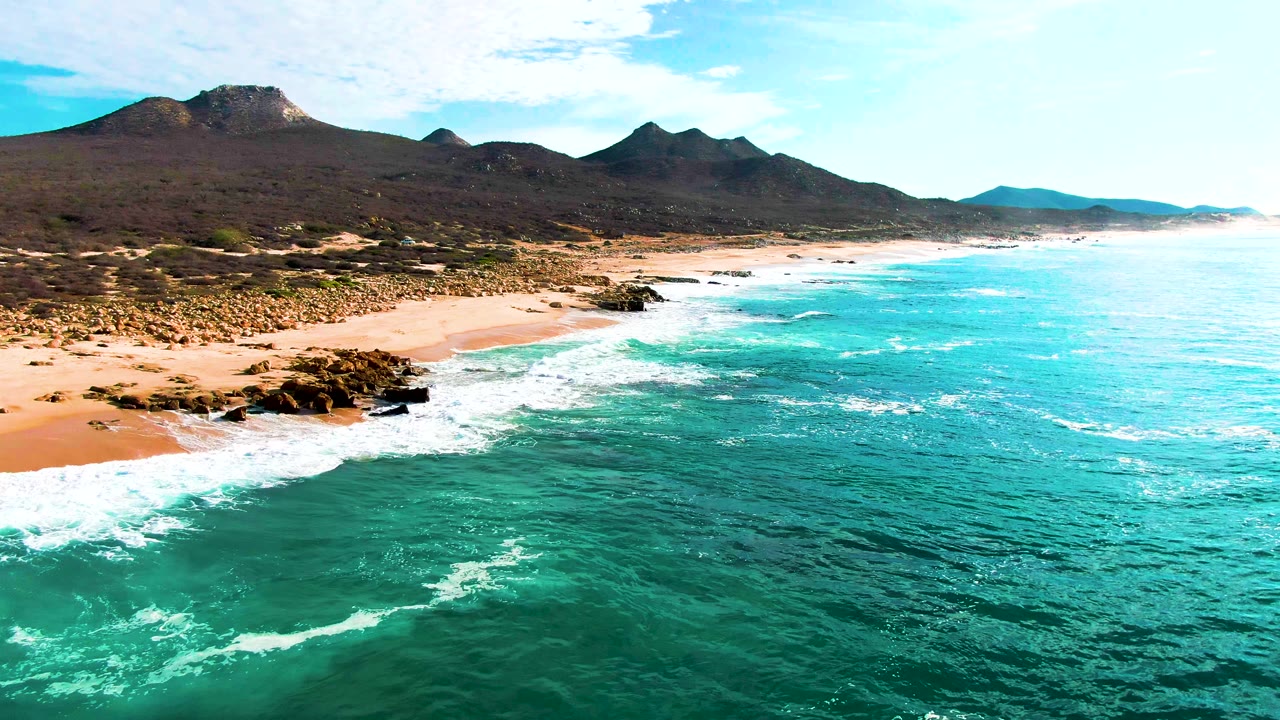 Aerial shoreline view with surf washing around rocks below dark mountains.
