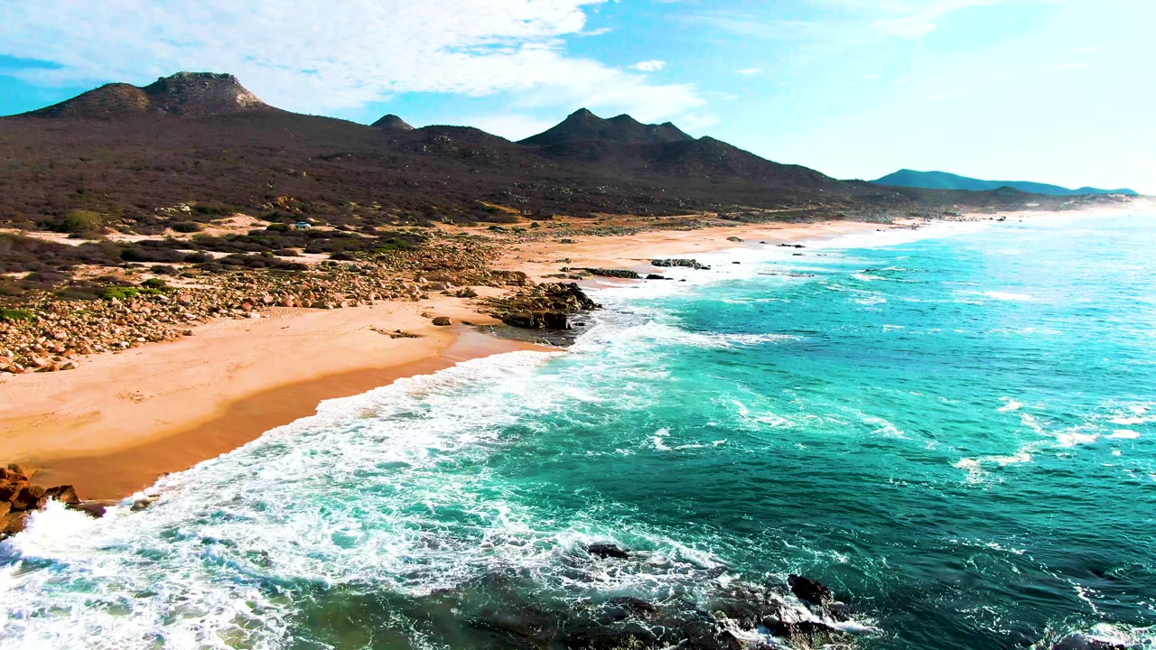 Aerial view of turquoise surf reaching a sandy beach below dark mountains.