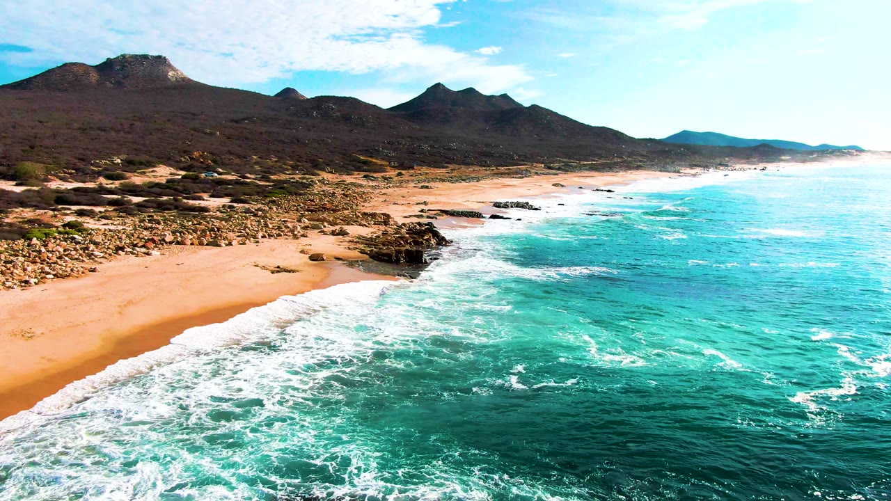 Aerial frame at Punta Buzzards showing white foam spreading farther across turquoise nearshore water, with sand and rocks on the left and mountains behind, no visible people.