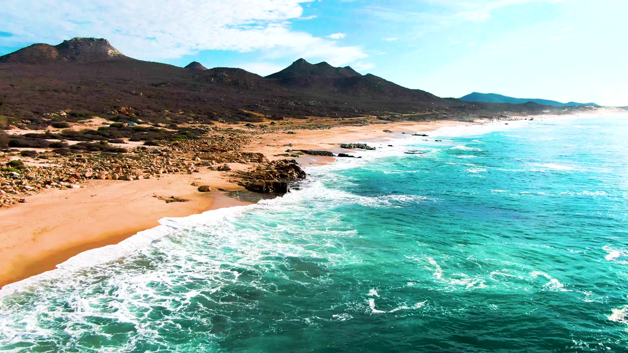 Aerial view along Punta Buzzards with a broader band of white surf across the right side, sandy beach curving left, scattered rocks near shore, and no visible people.