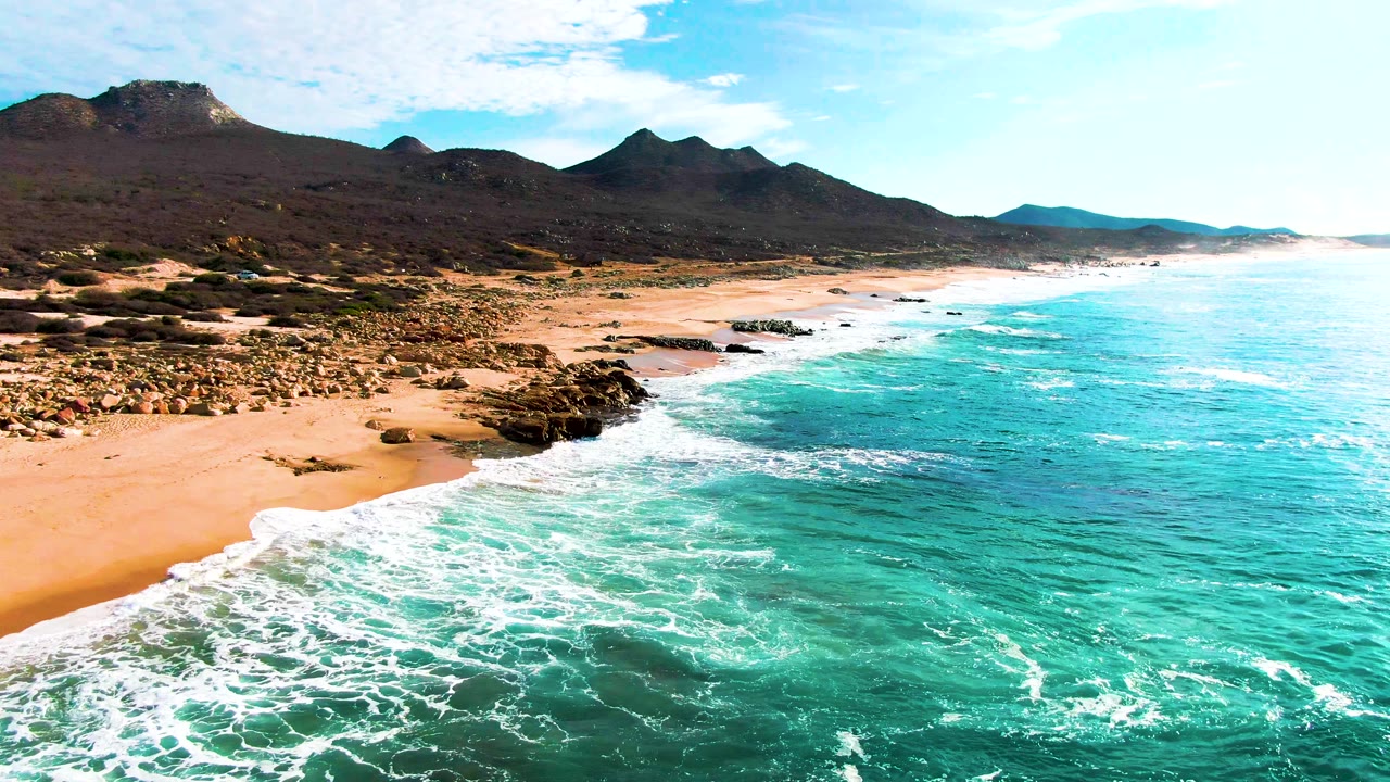 Mid-clip aerial frame over Punta Buzzards where the surf line runs diagonally across the foreground, dark rocks sit near the beach, mountains stretch across the horizon, and no visible people appear.