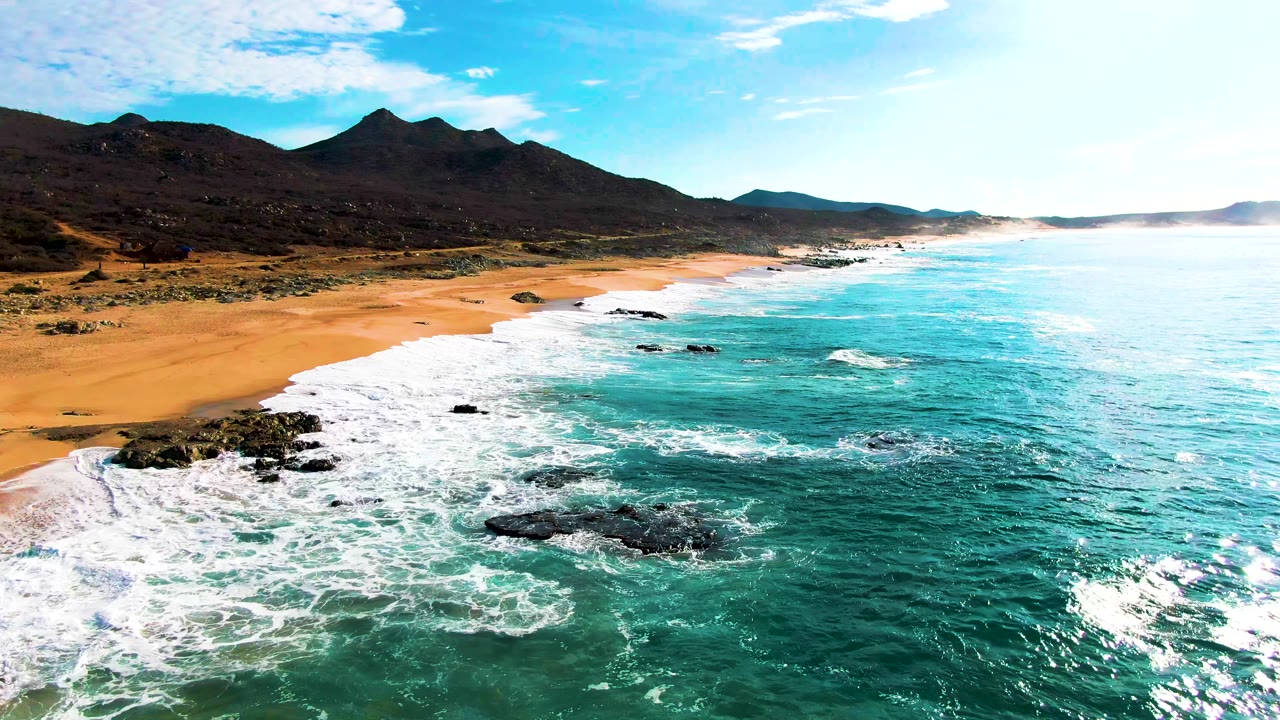 At Punta Buzzards in Baja California Sur, turquoise waves spread white foam across rocks and sand with dark mountains behind the beach and no visible people.