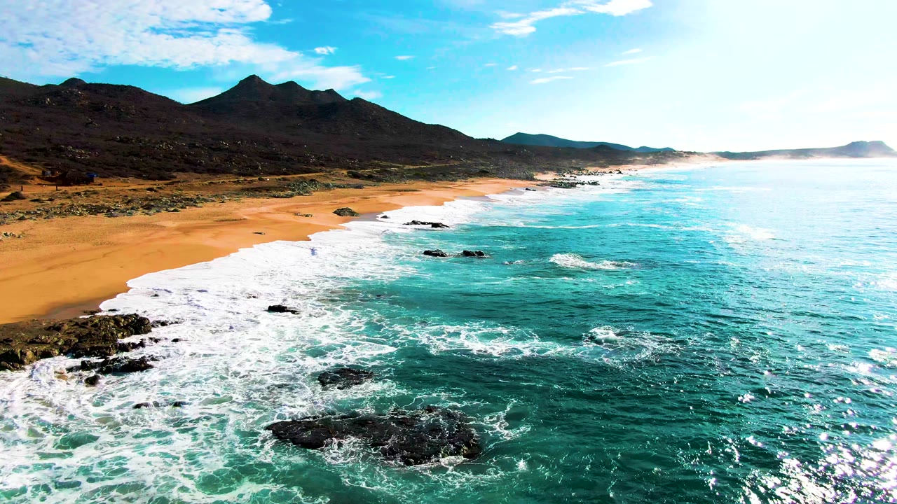 At 0:01, the Punta Buzzards shoreline runs diagonally through the frame as white surf curls around dark rocks, orange sand, and mountain slopes with no visible people.