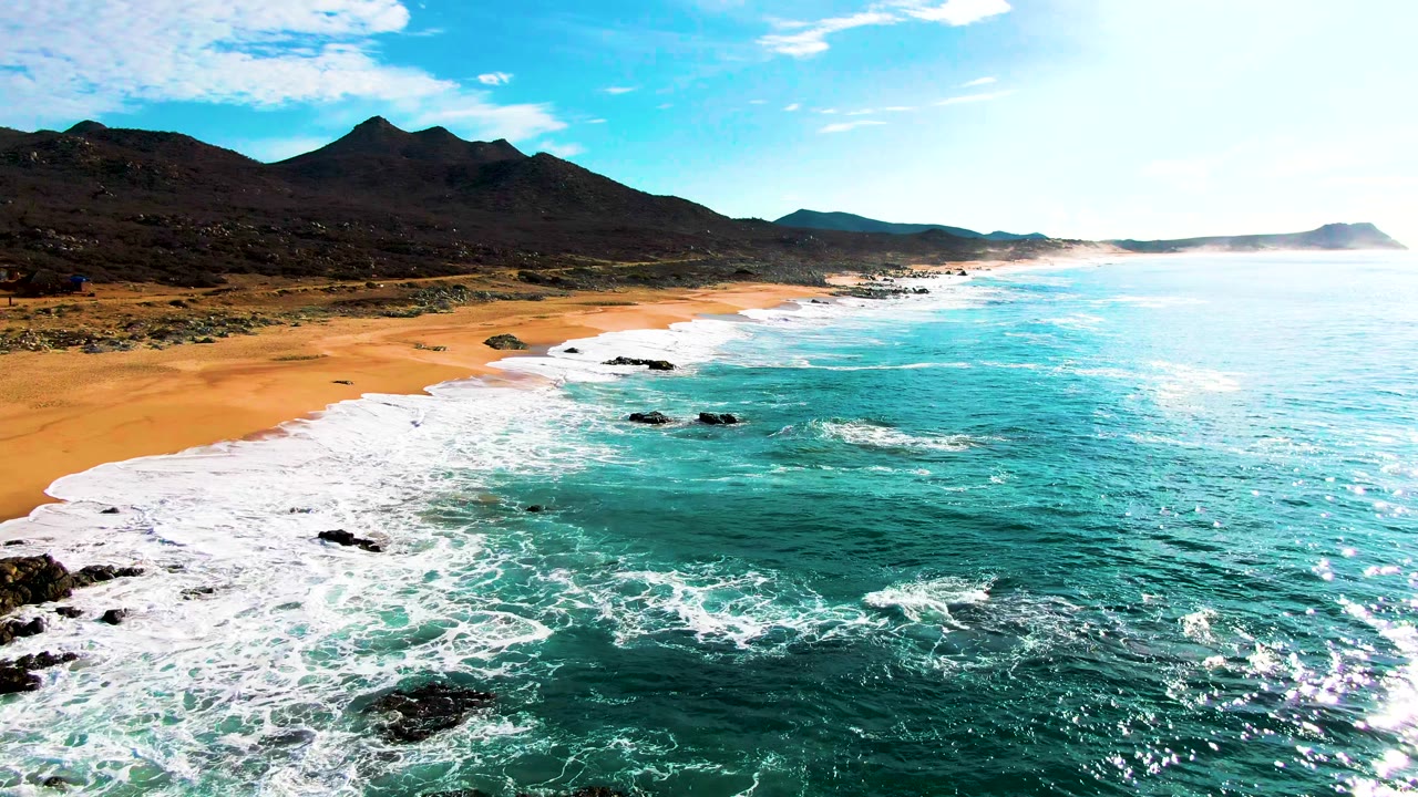At 0:02, bright glare spreads across the right side of the ocean while waves break across the sandy Punta Buzzards beach and no visible people appear onshore.