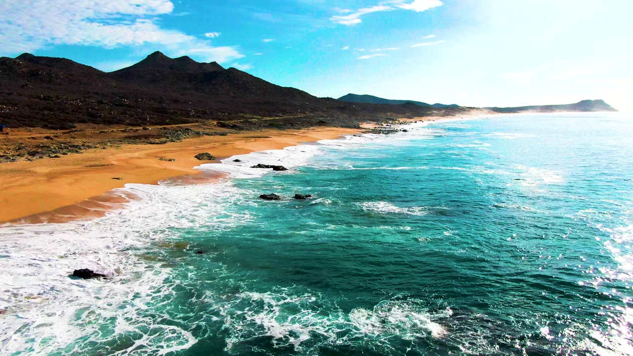 Aerial beach view with surf washing around dark rocks and low mountains behind.